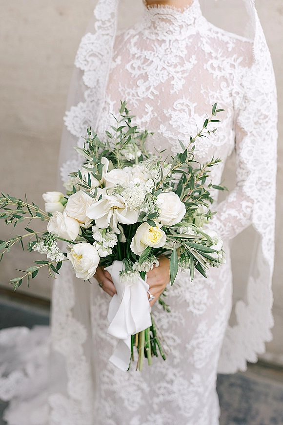 Bridal bouquet of white roses with greenery, tied with a ribbon, held against a lace wedding dress and veil near a neutral wall