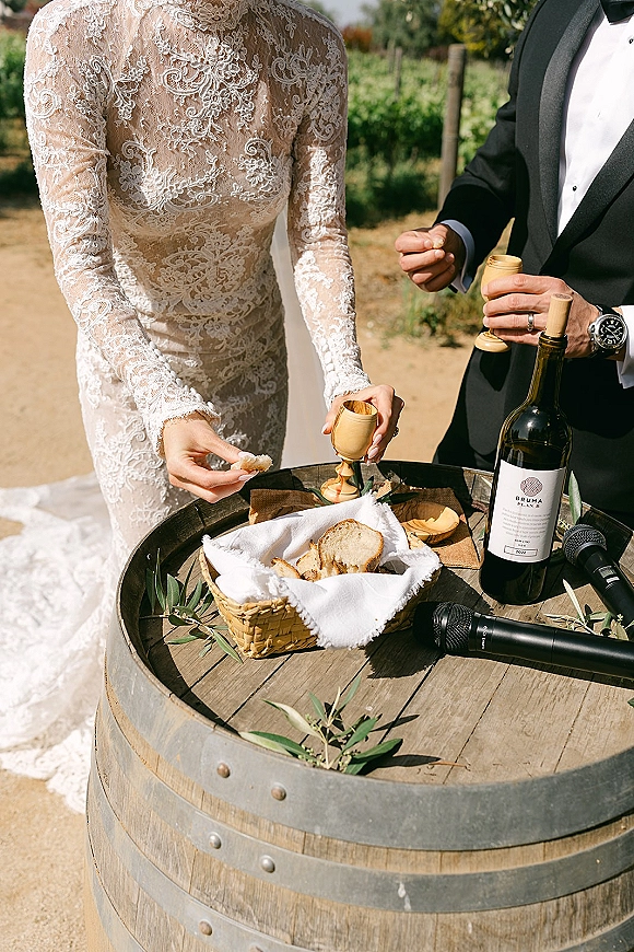 Wedding communion ceremony with bride in lace long sleeves and groom in tux sharing bread and wine at a barrel table in a vineyard