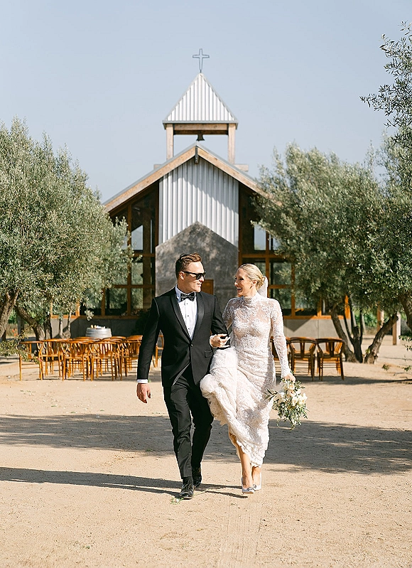 Wedding couple portrait of bride and groom walking down an outdoor aisle by a chapel with a cross, bride holding a bouquet, groom in sunglasses
