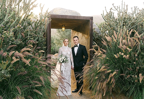 Couple portrait of bride and groom holding hands, bride in lace long sleeve dress with veil and white rose bouquet in desert doorway