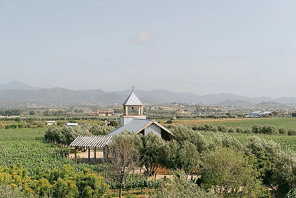 Wedding chapel set at a vineyard wedding venue with a bell tower and cross, wooden chairs, and vineyard rows with mountains beyond