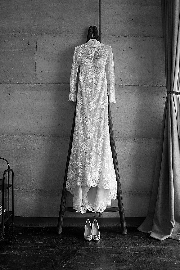 Wedding dress with long sleeve lace wedding dress details hanging on a wooden ladder, scalloped hem and bridal shoes against a concrete wall