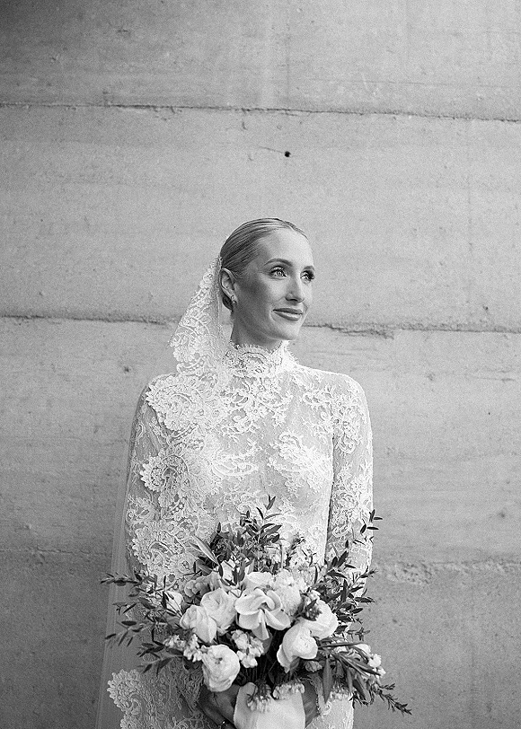 Bridal portrait in black and white of a bride looking away, holding a rose and greenery bouquet in a lace high-neck gown by a stone wall
