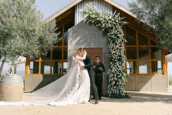 Wedding kiss under a white floral arch as the bride’s long veil falls behind, with an officiant at a modern glass venue courtyard
