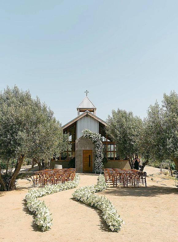 Ceremony setup with wood ceremony chairs and lush aisle florals leading to chapel double doors beneath a cross, with olive trees and blue sky