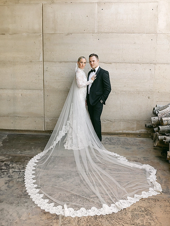 Couple portrait of bride in long sleeve lace wedding dress with cathedral veil beside groom in black tuxedo against concrete wall