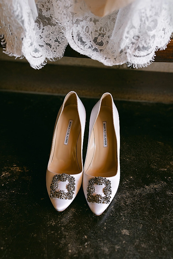 Bridal shoes in white satin wedding heels with crystal buckle clips beside a lace dress hem on a dark floor by a bench