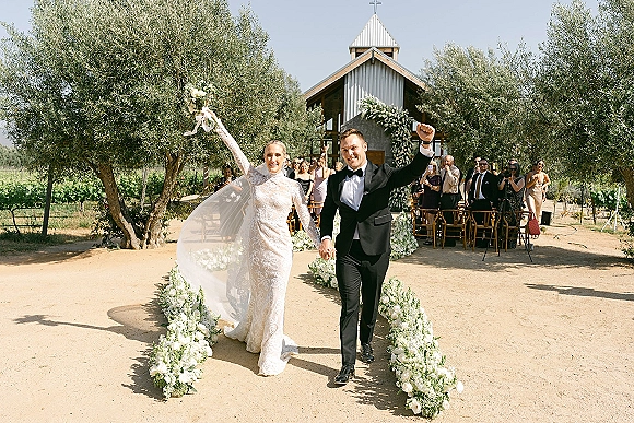 Wedding recessional as newlyweds walk down the aisle cheering, bride in lace long sleeves with veil and bouquet, guests by chapel vineyard rows