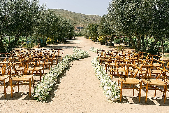 Ceremony aisle design with outdoor wedding ceremony setup featuring wood chairs and low white florals along a dirt path in an olive grove vineyard