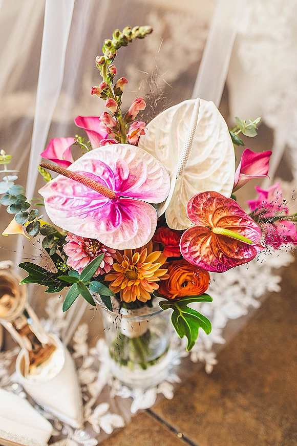 Bridal bouquet with tropical bridal bouquet blooms, anthurium and calla lilies beside white heels on lace fabric over a wood floor