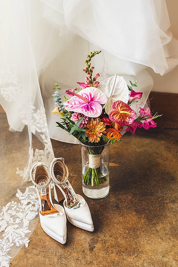 Wedding details flatlay with bridal bouquet and shoes, featuring colorful anthurium blooms, lace veil, and pink ribbon on stone floor