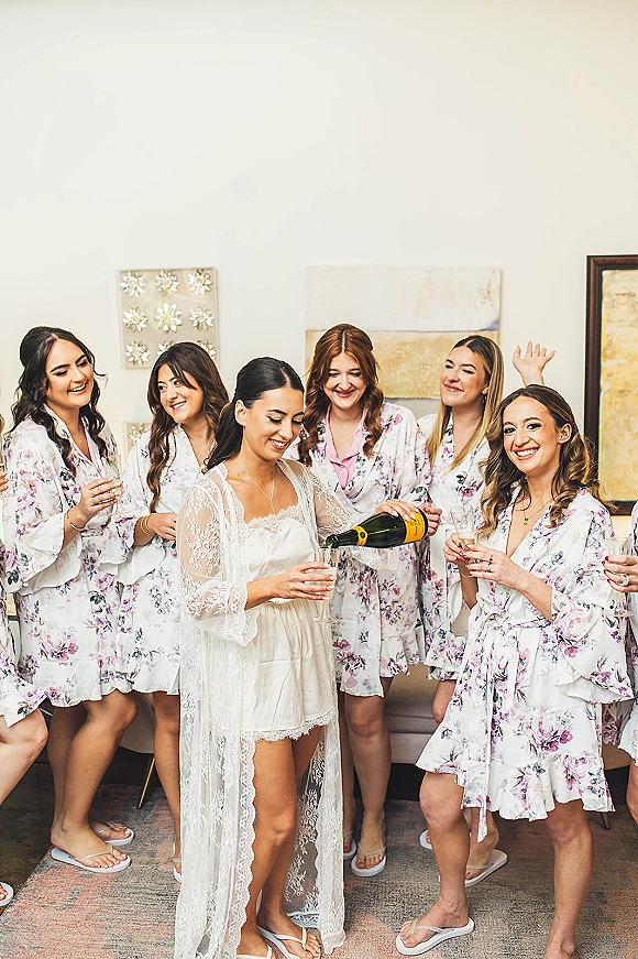 Bridal party getting ready in bride and bridesmaids robes, toasting champagne with flutes in an indoor suite with sofa and wall art
