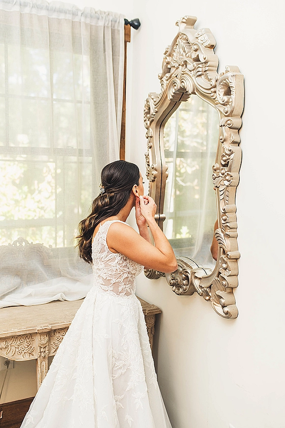 Bridal getting ready as bride putting on earrings in an ornate gold mirror, lace bodice visible beside a vanity in window light