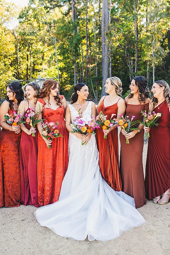 Bride with bridesmaids in rust dresses holding colorful bouquets, bride in white gown on a sunlit forest path with greenery