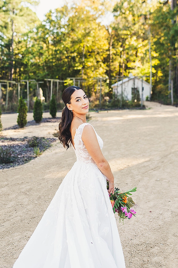 Bridal portrait of a bride looking back, holding a colorful bouquet in a lace wedding dress on a garden dirt path with trees