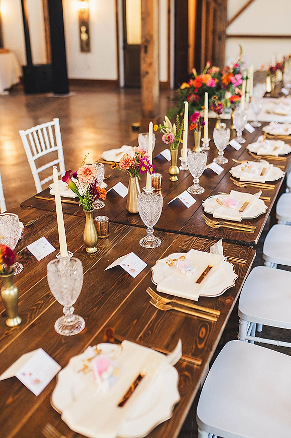 Reception tablescape on a farmhouse wedding table with bud vase florals, white taper candles, brass vases, and place cards in a barn interior