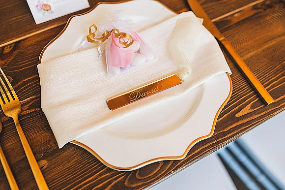 Wedding place setting with gold flatware, white plate on gold-rim charger, napkin ribbon curls, pink tulle almond favor on wood table