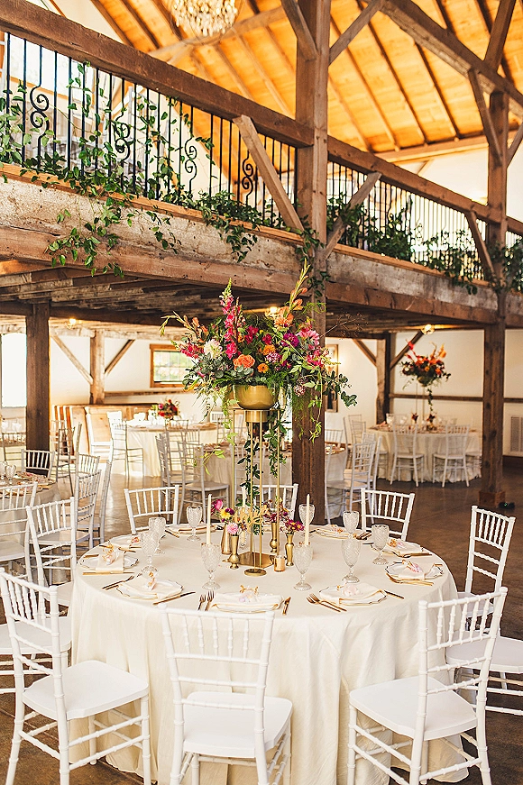 Reception tablescape with wedding reception table decor, tall floral centerpiece in a gold compote, taper candles, and greenery in a rustic barn interior