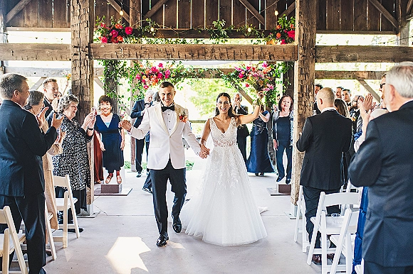 Wedding recessional as bride and groom walk the barn aisle hand in hand, bride raising her bouquet while guests cheer under string lights