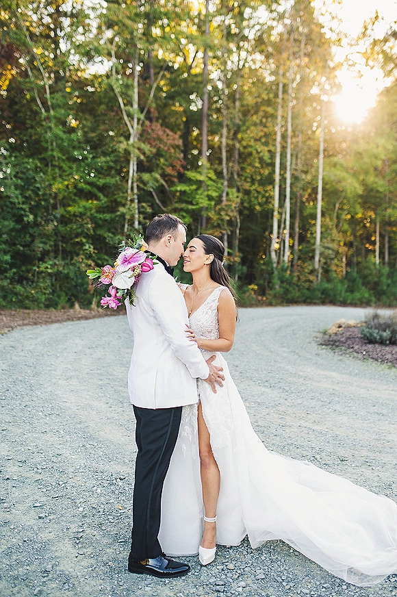 Couple portrait of bride and groom embrace on a sunlit gravel road, her bouquet and lace gown framed by forest trees