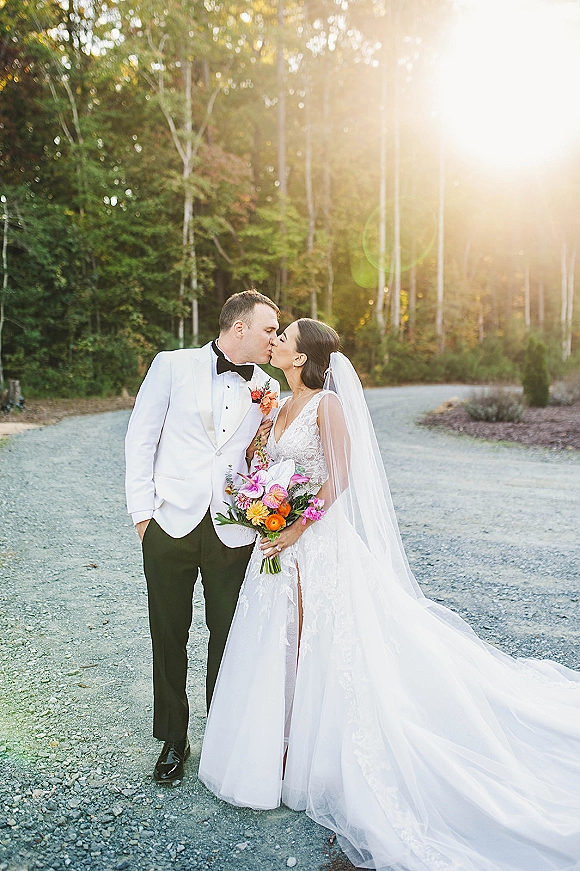 Wedding kiss portrait of bride and groom kissing on a sunlit gravel road in the forest, bride holding a colorful bouquet and veil flowing