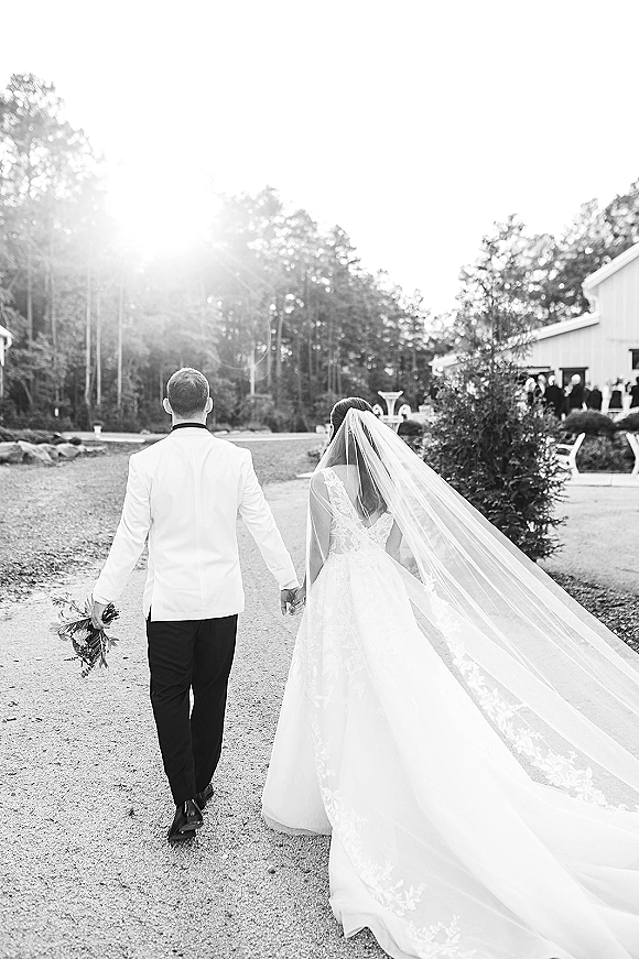 Wedding couple walking hand in hand from behind, bride’s long veil and lace train flowing with bouquet on a sunlit gravel path near a barn