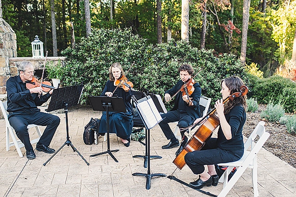 Wedding string quartet in black attire plays violins and cello from sheet music on a patio beside a stone fireplace and garden greenery