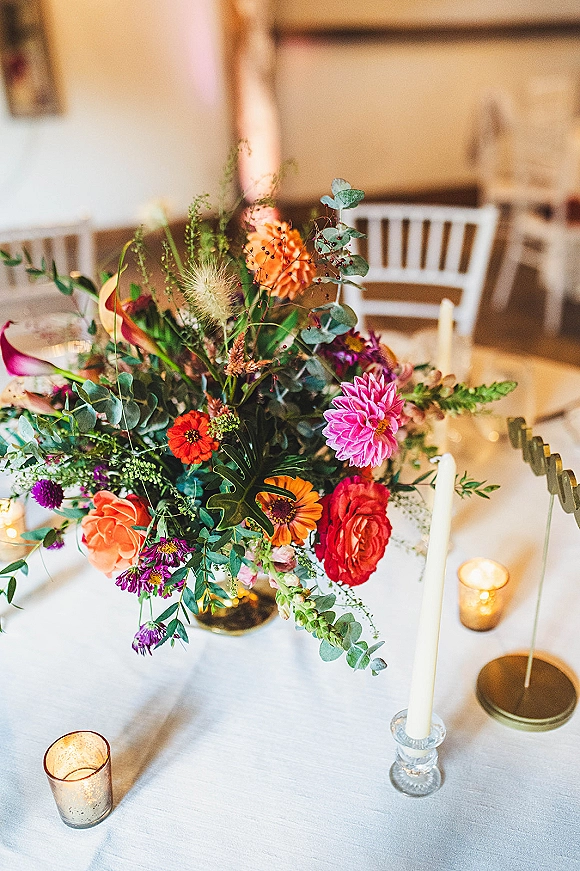 Reception centerpiece with wildflower wedding centerpiece blooms, eucalyptus and white taper candles in glass holders on a white tablecloth in reception room