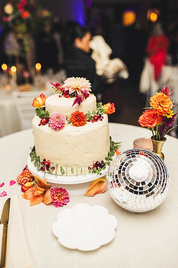 Wedding cake with buttercream frosting topped with fresh roses and dahlias, accented by a disco ball on a stand in a bokeh-lit reception room