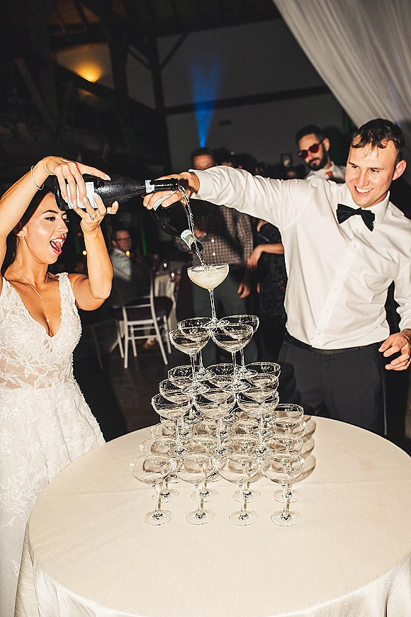 Champagne tower at an indoor reception as bride and groom pour bubbly into coupe glasses on a white table under bistro lights