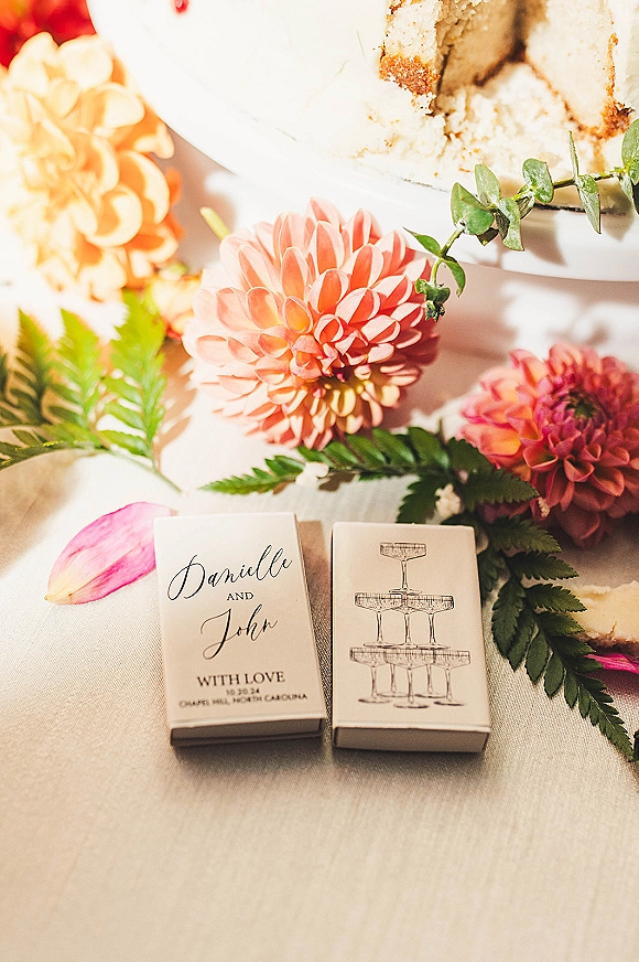 Wedding matchboxes and custom wedding matchbooks with calligraphy and illustration beside a white cake, dahlias, and fern greenery on linen.