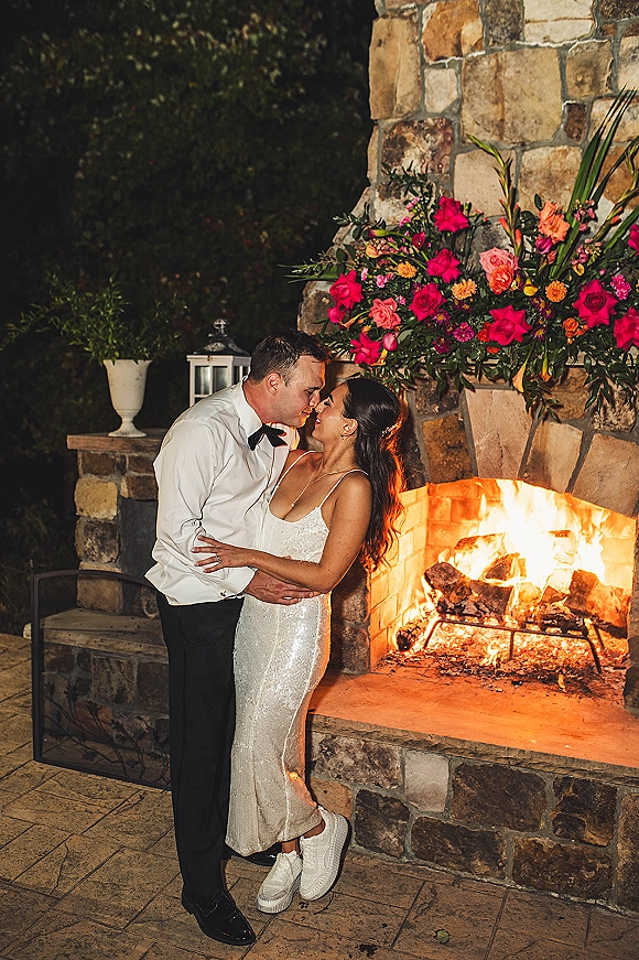 Wedding kiss portrait of bride and groom kissing in a sparkly gown and bow tie beside an outdoor fireplace with lantern glow at night