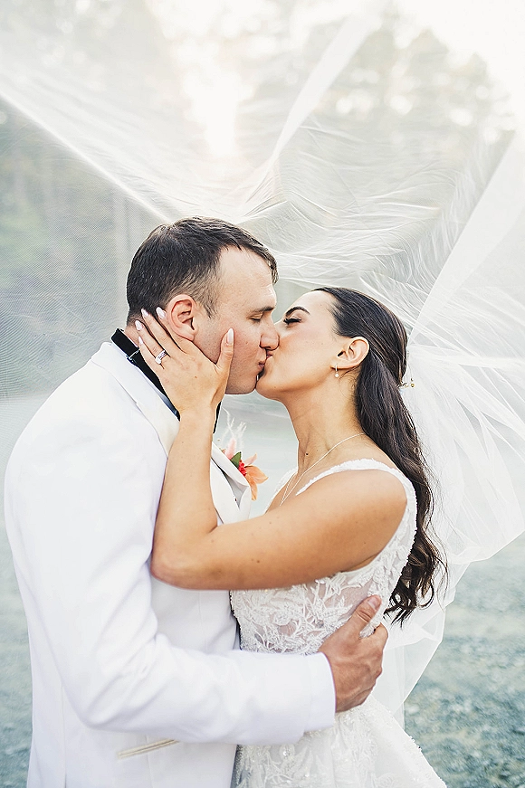 Wedding kiss portrait of bride and groom kissing as her veil blows around them, her hand on his face, outdoors with soft greenery bokeh