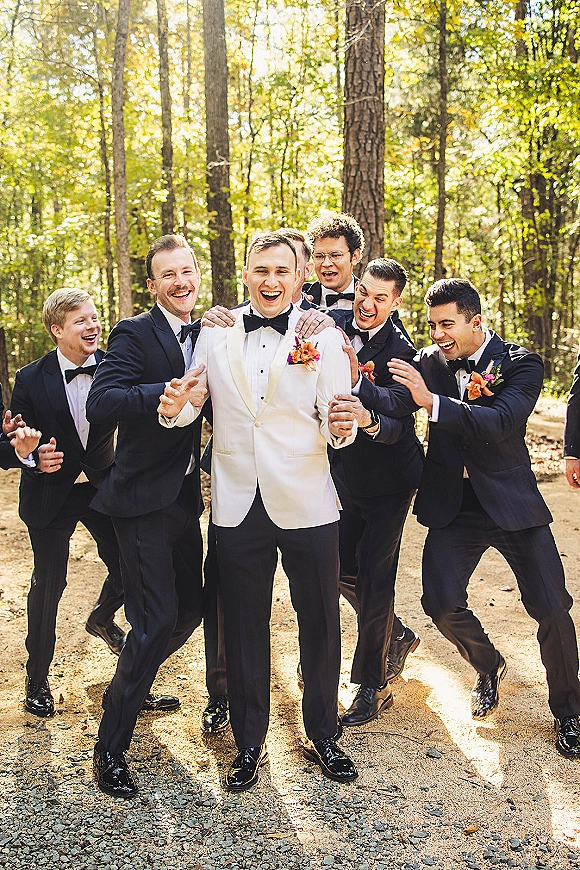 Groomsmen portrait of groom with groomsmen laughing in tuxedos, groom in white dinner jacket, on a sunlit forest path with trees