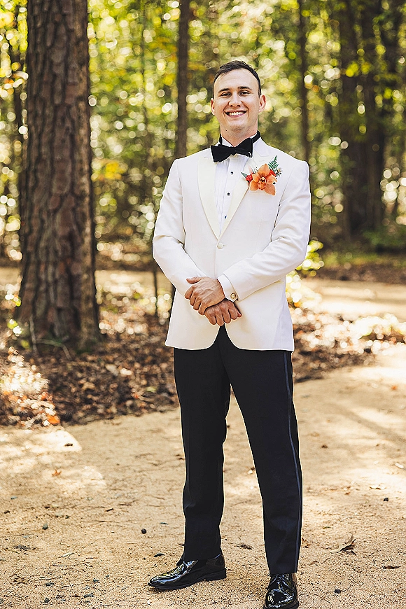 Groom portrait in a white tuxedo jacket with black bow tie and boutonniere, standing on a forest dirt path amid trees and greenery
