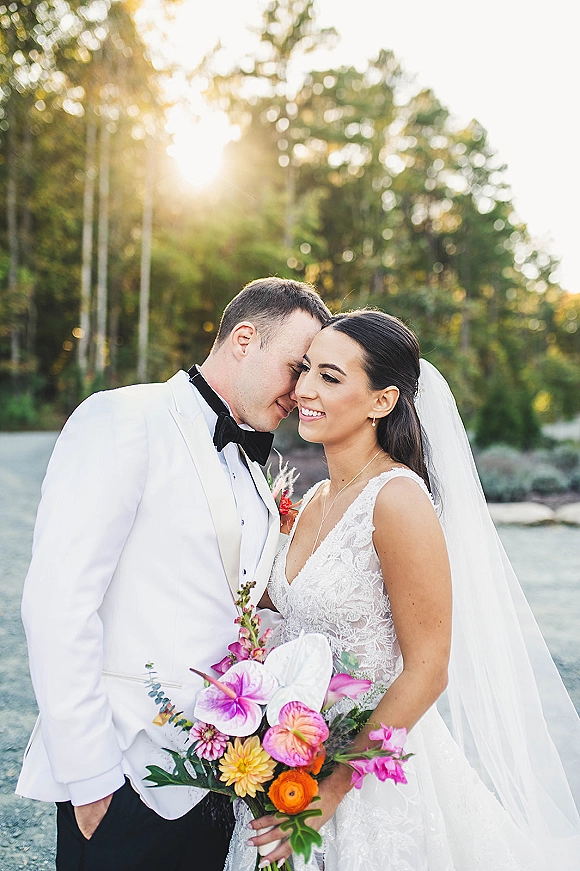 Couple portrait of bride and groom in close embrace, foreheads touching, backlit in forest trees on a gravel road with sun flare