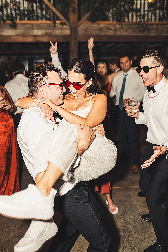 Wedding dance moment as groom lifts bride in heart sunglasses, cocktail glass raised, glow necklace shining under string lights and wooden beams