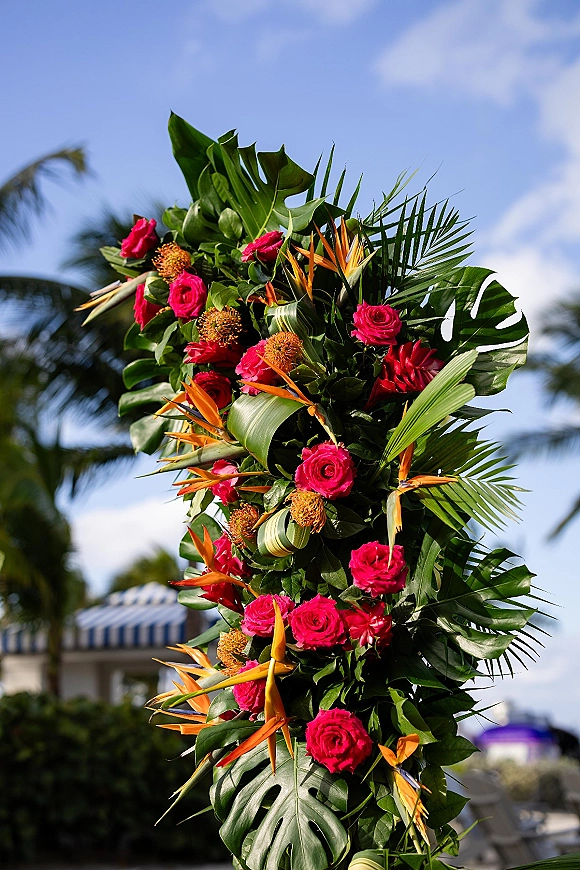 Tropical wedding florals on a tall ceremony arrangement with bird of paradise, hot pink roses, and monstera leaves beneath blue sky and palms