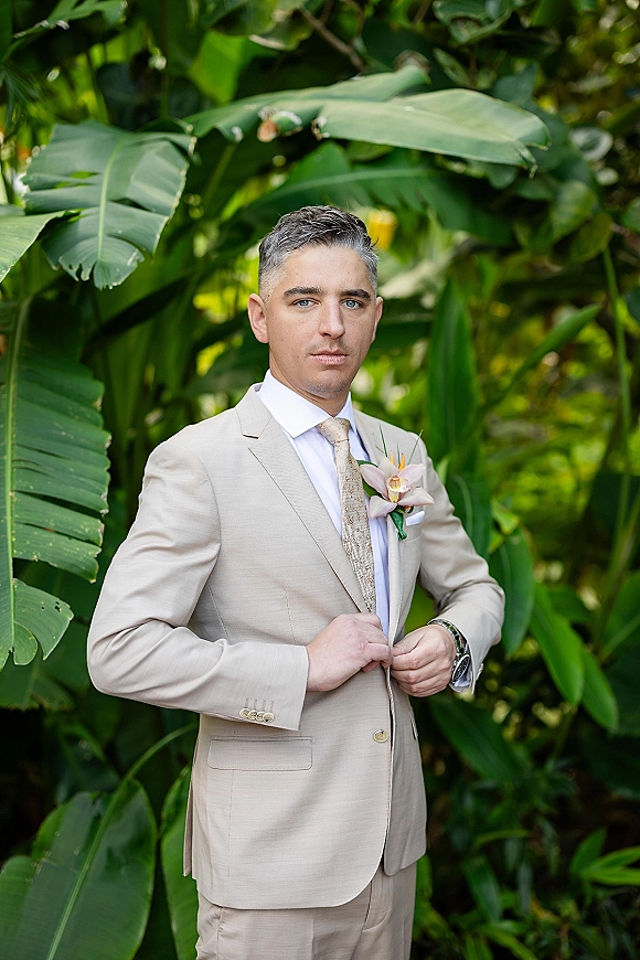 Groom portrait in a beige groom suit adjusting his jacket, wearing a patterned tie, orchid boutonniere, and wristwatch amid lush tropical foliage