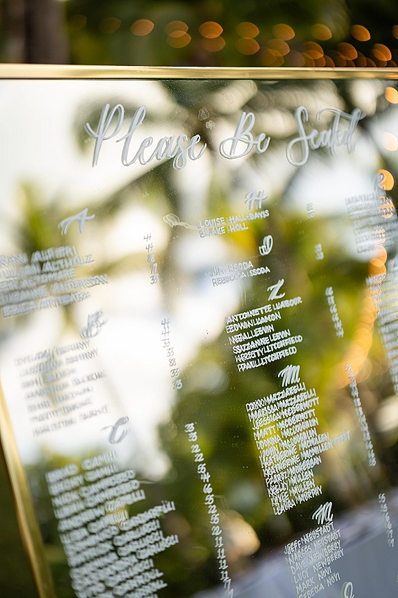 Wedding seating chart on a gold-framed mirror seating chart with white calligraphy lettering, set outdoors amid greenery and bokeh lights