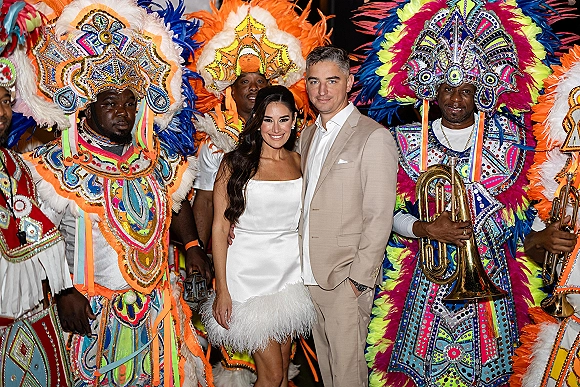 Wedding couple portrait with bride in strapless satin feather-hem mini dress and groom in tan suit beside brass band in a nighttime crowd