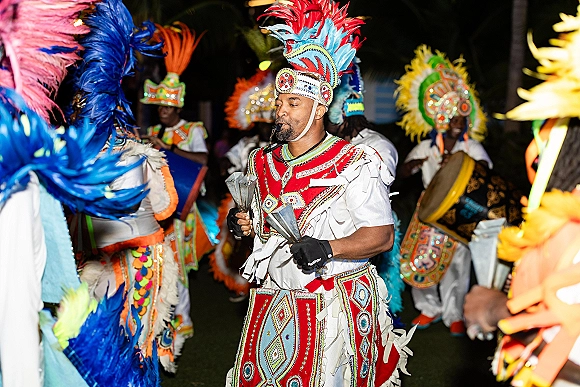 Wedding reception dancers in feather headdresses lead a wedding cultural dance with drums and whistles on a palm-lined lawn at night
