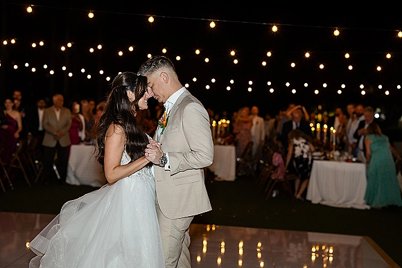 First dance as bride in a strapless wedding dress and groom in a suit sway on a white dance floor under string lights, guests and candles behind