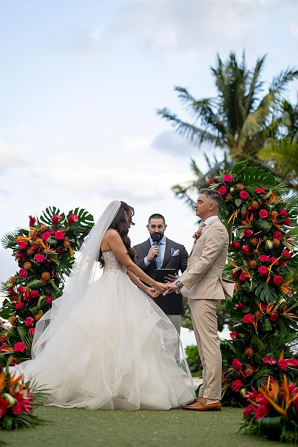 Wedding vows as bride in ball gown and groom in tan suit hold hands under a tropical arch with pink roses, palm trees behind