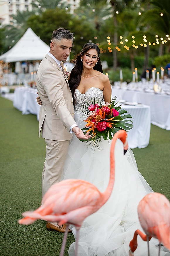 Couple portrait of bride and groom posing, she holds a tropical bridal bouquet under string lights with palm trees and a white tent behind