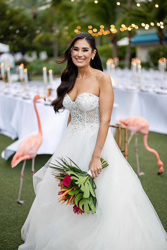 Bridal portrait of a smiling bride in a strapless lace wedding dress holding a tropical bouquet at palm-lit outdoor reception tables with string lights