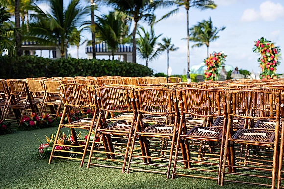 Ceremony seating with bamboo folding chairs lined along a greenery garland aisle, leading to a tropical flower arch under palm trees