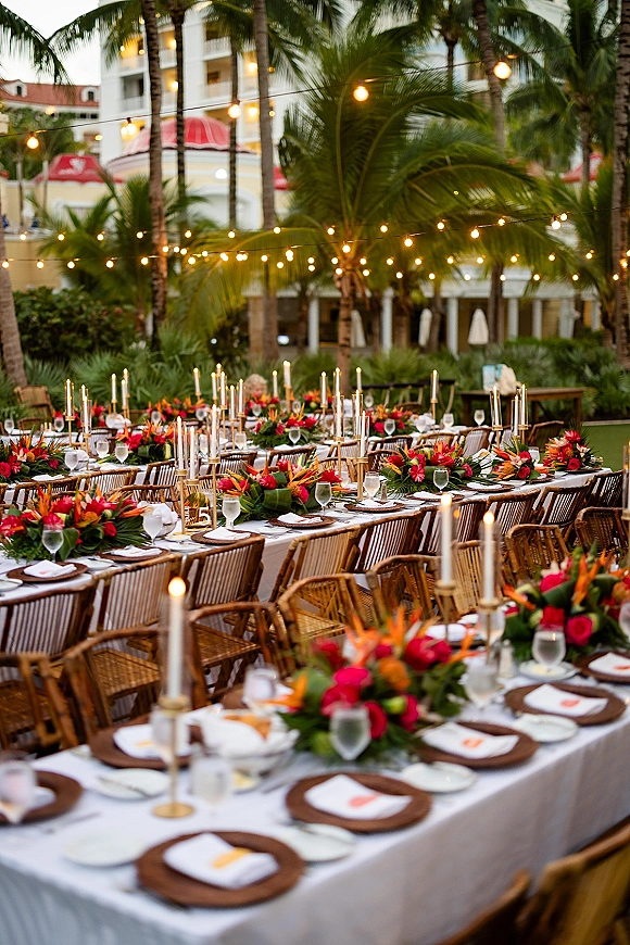 Reception tablescape at an outdoor wedding reception with long banquet tables, tropical floral centerpieces, taper candles, and palm trees under string lights