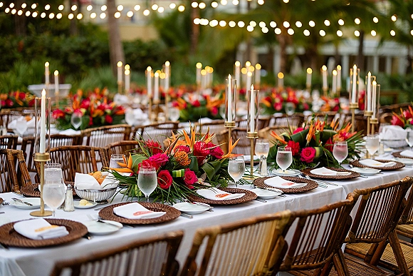 Reception tablescape with tropical wedding tablescape florals, taper candles in brass holders, white linens and rattan placemats under string lights in a palm courtyard