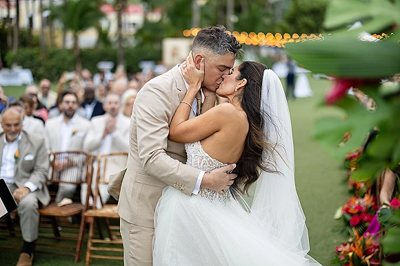Wedding kiss as bride in strapless lace dress and veil holds groom in beige suit, with guests, chairs, string lights, and tropical florals outdoors